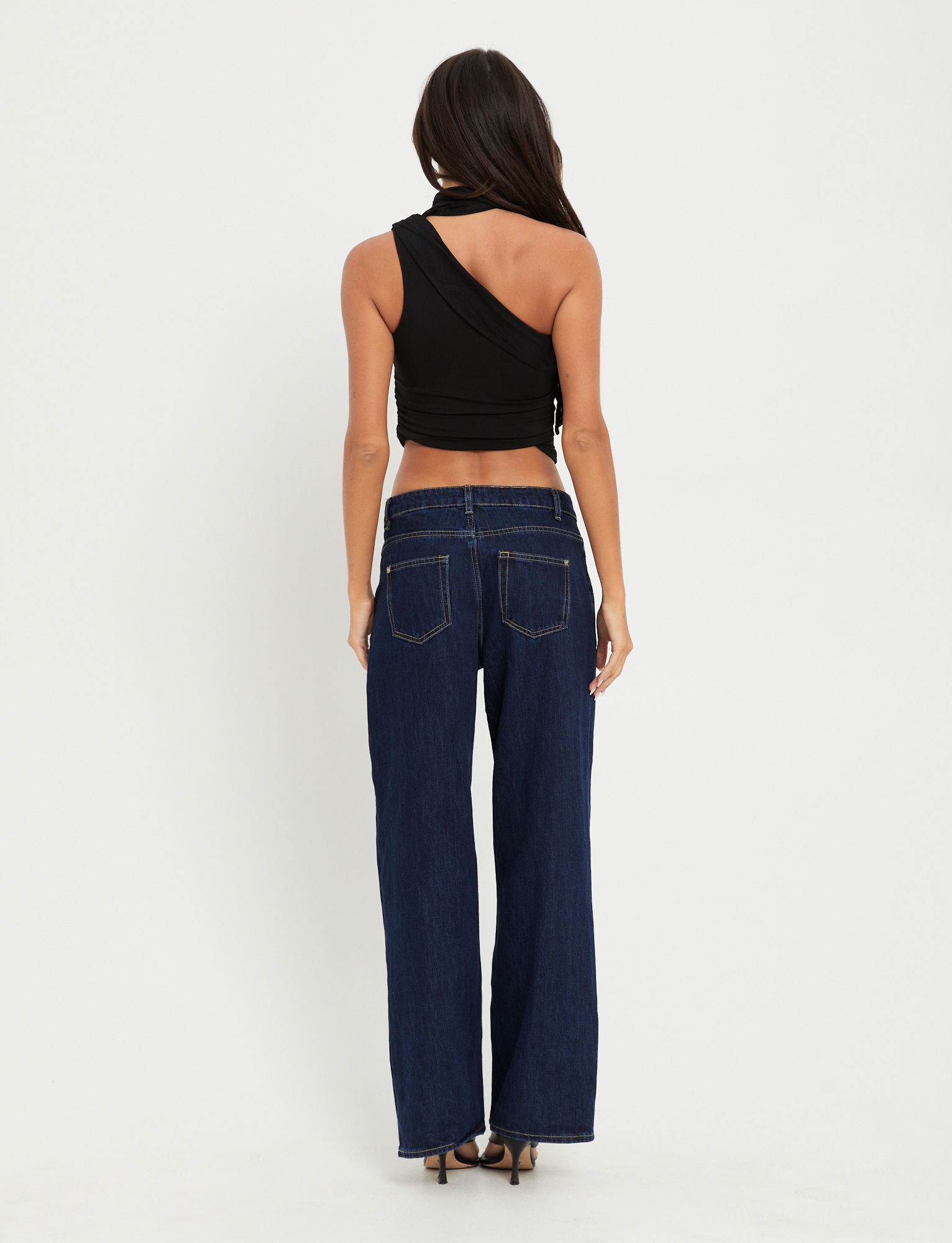 A woman faces away, wearing the TIGERMIST ALLIRA TOP in black— a high-stretch, one-shoulder tank—paired with dark blue wide-leg jeans and high heels against a plain white background.