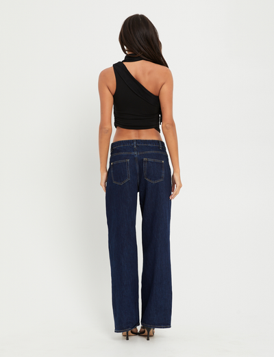 A woman faces away, wearing the TIGERMIST ALLIRA TOP in black— a high-stretch, one-shoulder tank—paired with dark blue wide-leg jeans and high heels against a plain white background.