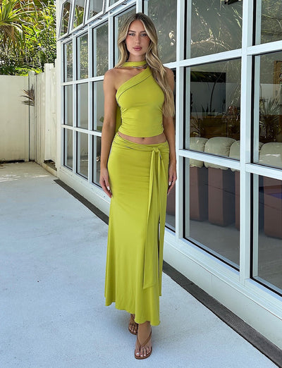 A woman stands in front of a glass wall wearing the ALLIRA TOP - GREEN, an olive green chartreuse one-shoulder crop top from TIGERMIST, paired with a matching long skirt featuring a tied waist detail. The ensemble is complemented by brown strappy sandals. She poses with a relaxed expression against a backdrop of greenery.