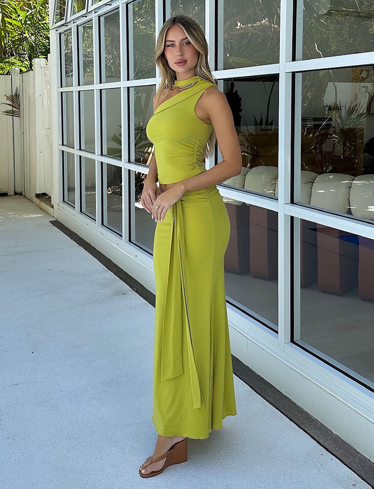 A woman wearing a chartreuse one-shoulder "MIST SKIRT - GREEN" dress from TIGERMIST stands on a patio next to a building with large windows. She has long hair and is looking towards the camera, while also sporting brown wedge sandals. The background features potted plants and reflections in the windows.