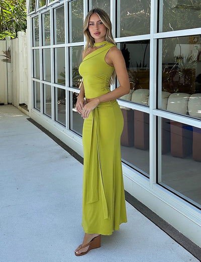 A woman wearing a chartreuse one-shoulder "MIST SKIRT - GREEN" dress from TIGERMIST stands on a patio next to a building with large windows. She has long hair and is looking towards the camera, while also sporting brown wedge sandals. The background features potted plants and reflections in the windows.