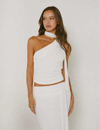 A woman with long, wavy brown hair is wearing the ALLIRA TOP - WHITE and a matching skirt from TIGERMIST. She stands against a plain background, looking to the side. Her look is completed with rings on her fingers.