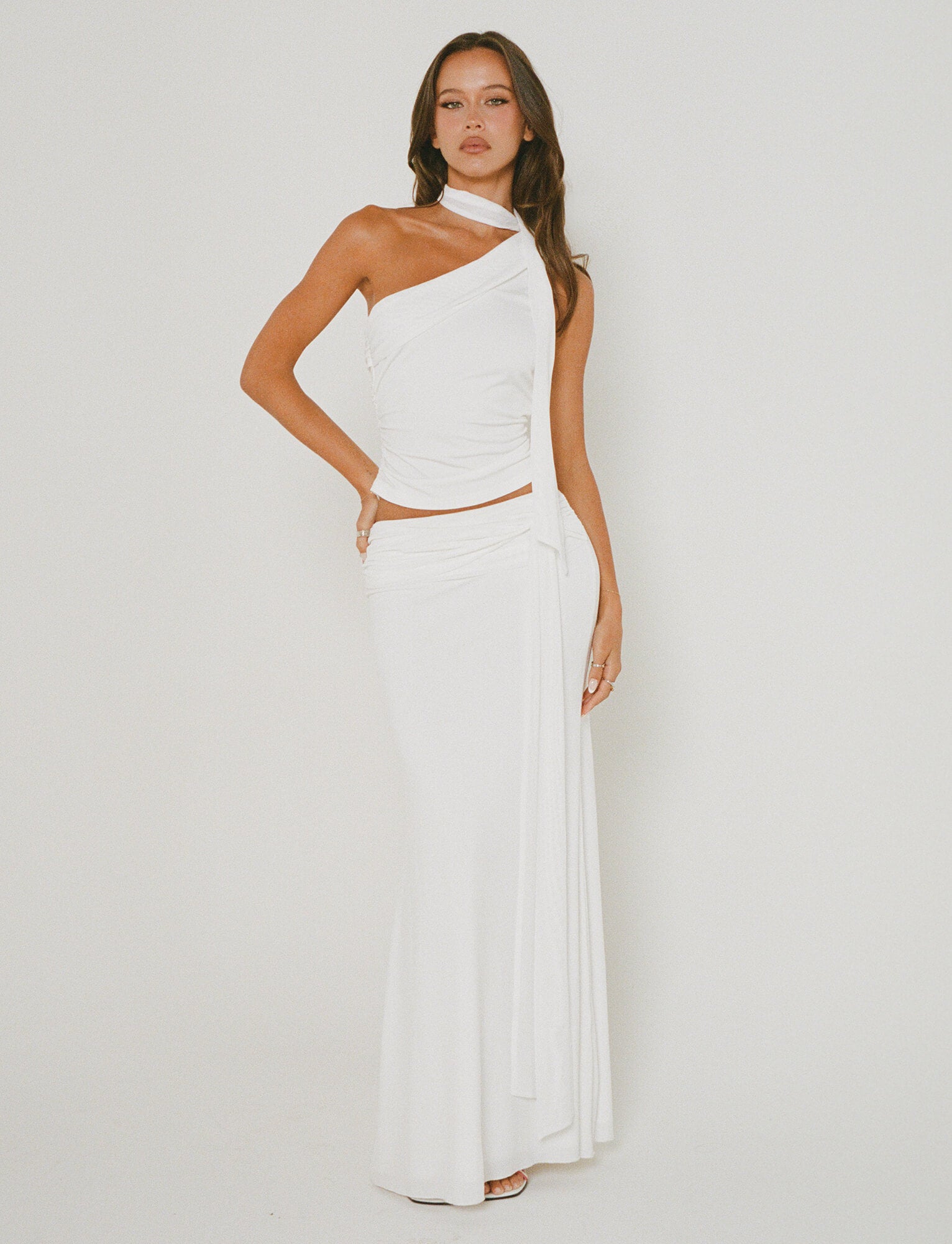 A woman stands against a plain background, wearing the stylish MIST SKIRT - WHITE from TIGERMIST. The skirt showcases an elegant wrap design with draped fabric, paired perfectly to highlight its chic one-shoulder style. She poses with one hand on her hip and the other resting by her side.