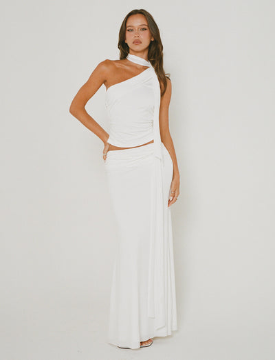 A woman stands against a plain background, wearing the stylish MIST SKIRT - WHITE from TIGERMIST. The skirt showcases an elegant wrap design with draped fabric, paired perfectly to highlight its chic one-shoulder style. She poses with one hand on her hip and the other resting by her side.