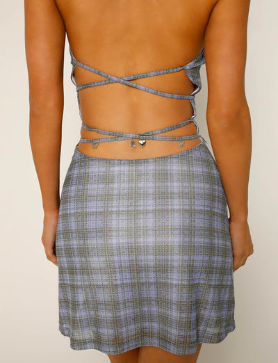 A woman shown from behind is wearing the PATRA DRESS - MULTI : MIRO CHECK by TIGERMIST. The dress features an open back with thin, crisscrossing straps and a small silver heart embellishment at the waist.