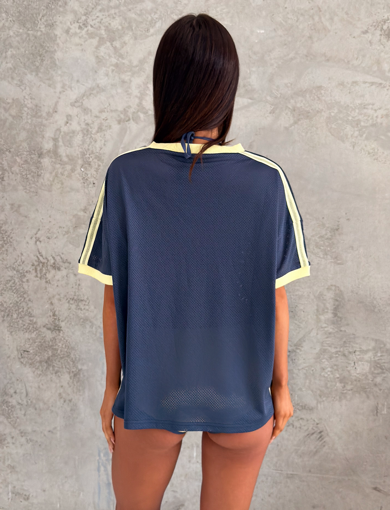 A person with long dark hair faces a textured gray wall, wearing the TIGERMIST JAYNA TEE in dusty blue—an oversized sheer mesh jersey t-shirt with yellow trim and no visible pants.
