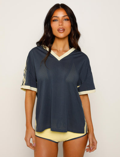 A person with long, dark hair is wearing a loose-fitting, dusty blue mesh V-neck shirt with light yellow trim and short sleeves from TigerMist's Jayna Tee collection. They are also wearing matching light yellow shorts. The background is plain and light-colored.