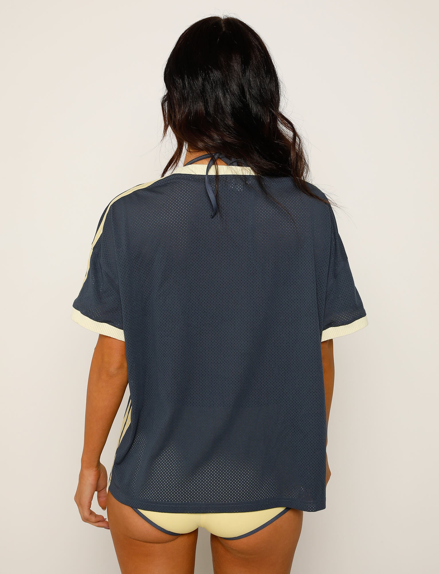 A person with long dark hair is standing with their back to the camera, wearing the TIGERMIST JAYNA TEE in dusty blue, featuring light yellow trim, along with matching light yellow bikini bottoms. The background showcases a plain, light-colored wall.