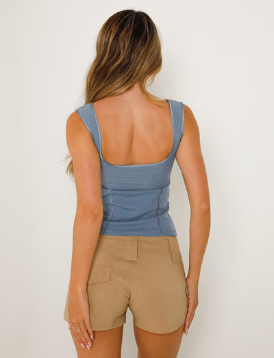 A person with long, wavy hair is facing away from the camera. They are wearing a sleeveless dusty blue top with a square back neckline, identified as the NOEMI TOP - BLUE from the stylish TIGERMIST collection, paired with beige shorts featuring pockets. The background is plain and off-white.