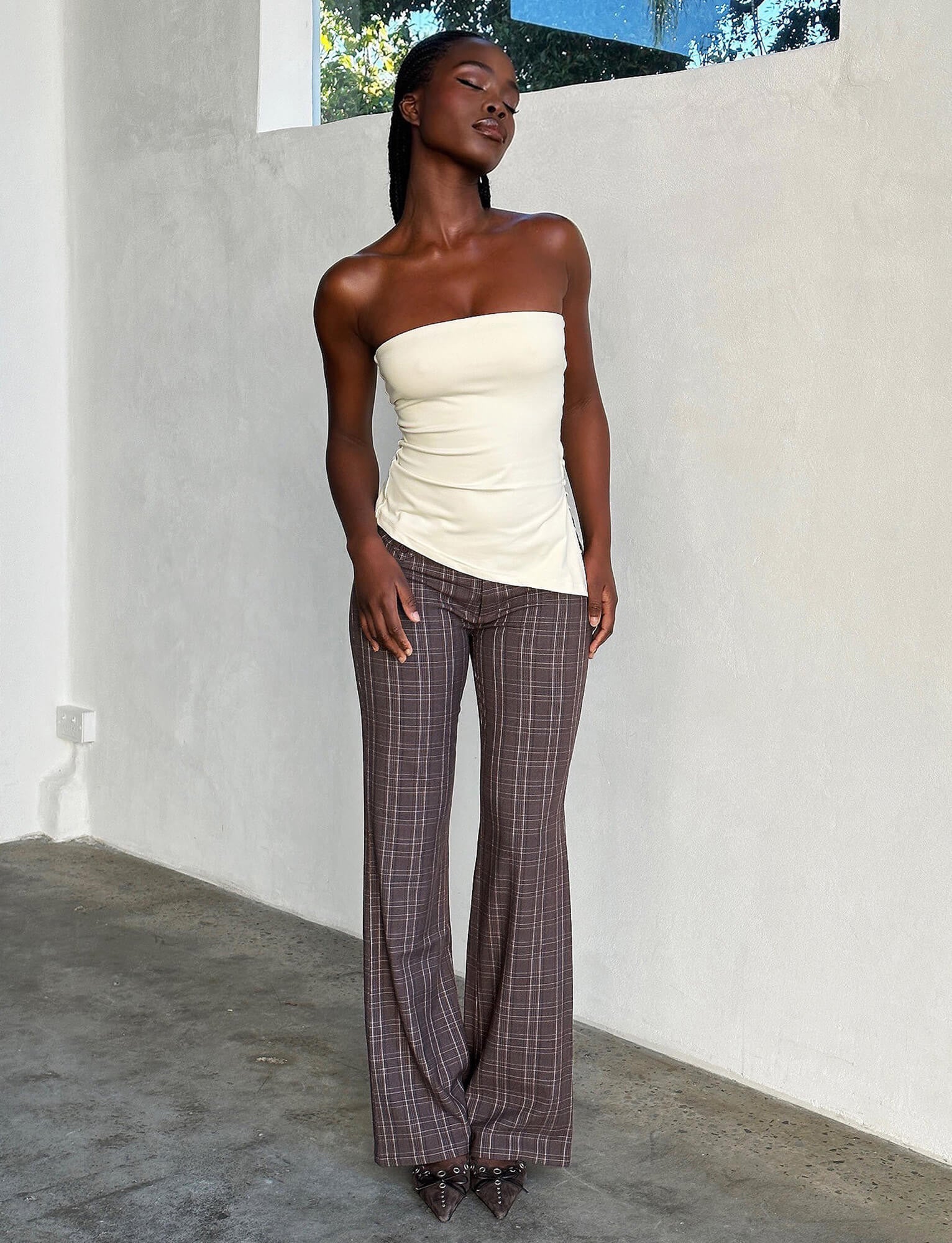 A woman stands in a minimalistic, well-lit space, donning a strapless, off-white top and KITTIE PANT - BROWN : CHOCOLATE CHECK from TIGERMIST, paired with pointed shoes. The room boasts a concrete floor and a plain wall with a window that offers a glimpse of trees outside.