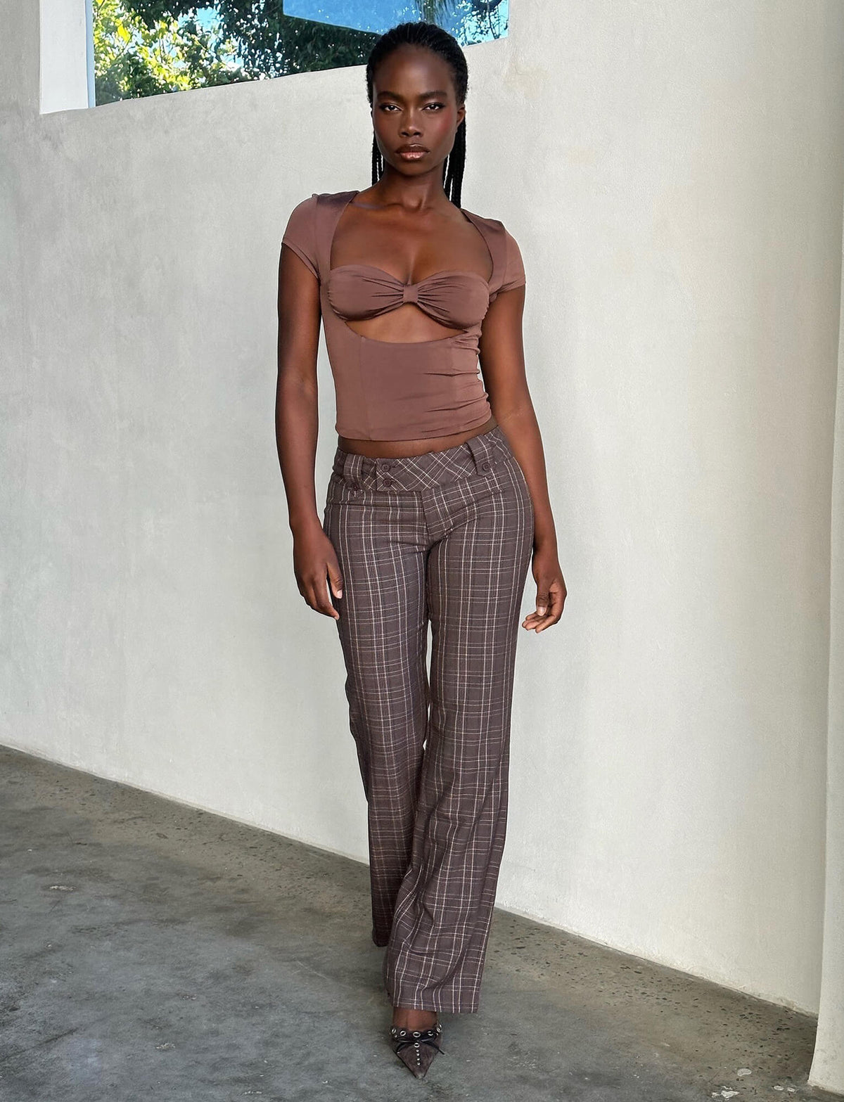 A woman with long black hair stands confidently indoors. She is wearing a fitted, short-sleeved brown top from Tiger Mist with a knotted detail in the front and the KITTIE PANT - BROWN : CHOCOLATE CHECK, high-waisted plaid pants from TIGERMIST. She completes the look with pointed-toe heels featuring intricate designs.