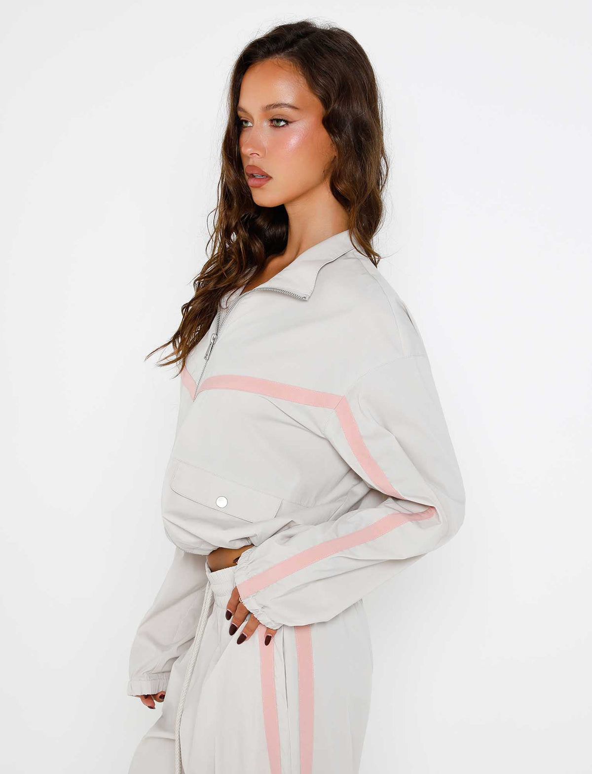 A person with long, wavy brown hair stands against a white background, sporting Tiger Mist's Dove Grey Kahanna Jacket with pink stripes on the sleeves and pants. They face slightly to the side, hands lightly held together in front.