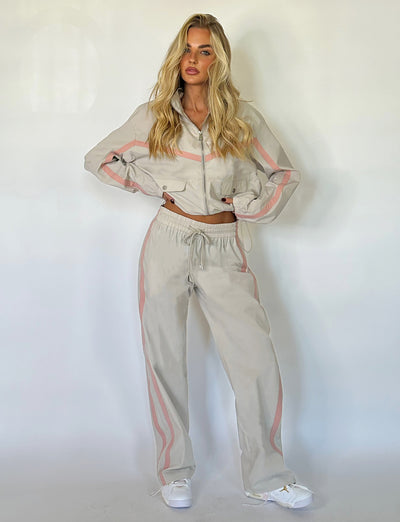 A person with long blonde hair stands against a plain white background, donning the TIGERMIST KAHANNA PANT in dove grey. The light beige tracksuit with pink stripes features a cropped jacket and high-waisted pants. The subject has their hands in their pockets and pairs the ensemble with white sneakers.