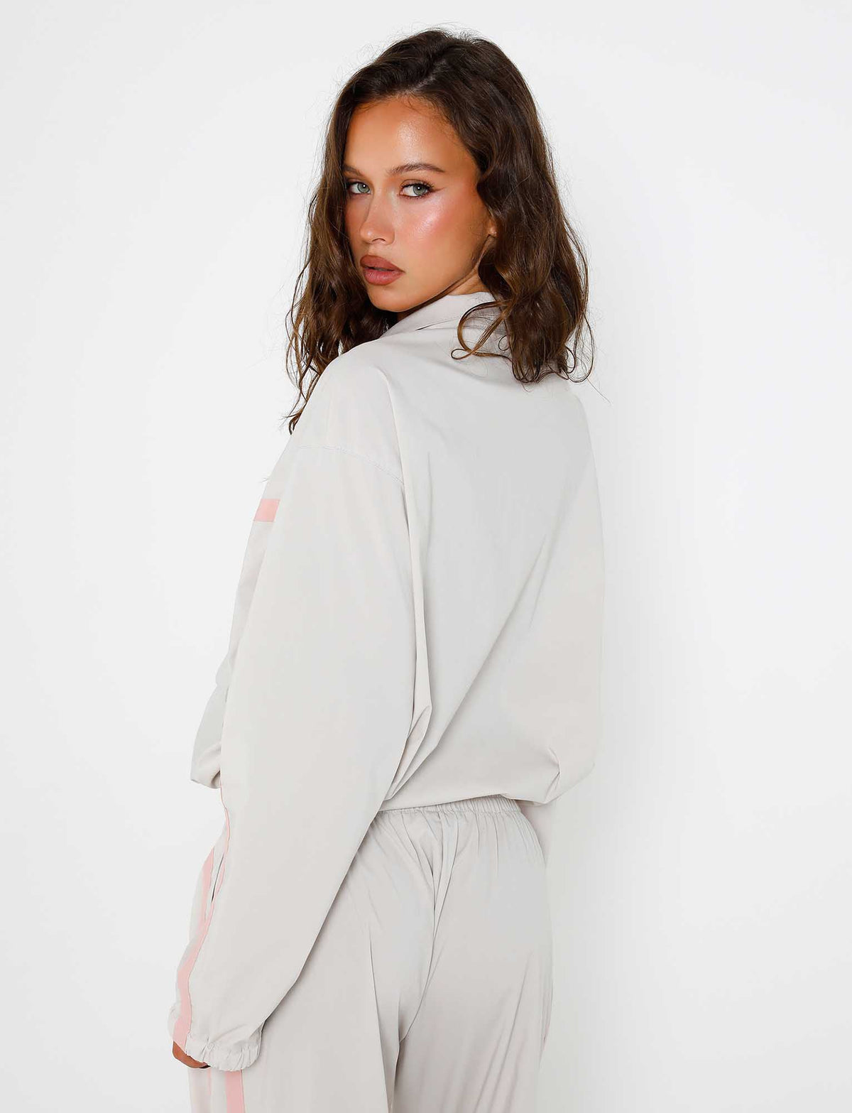 A person with wavy brown hair is looking over their shoulder towards the camera, dressed in a stylish TIGERMIST DOVE GREY KAHANNA JACKET and matching pants with a hint of pink detailing. The background is plain white.