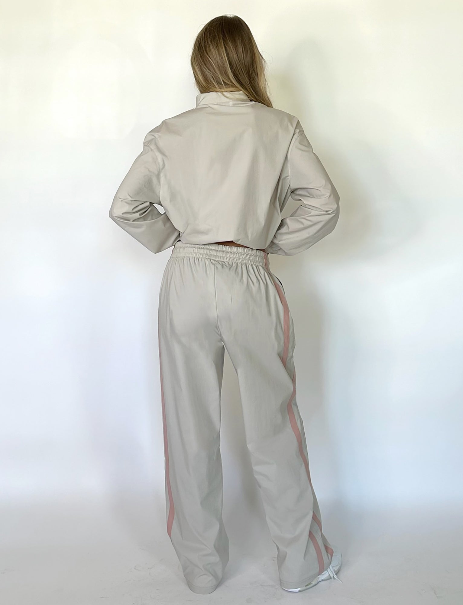 A person with long hair stands with their back to the camera, dressed in a chic TIGERMIST KAHANNA PANT in Dove Grey, featuring stylish pink stripes down the sides of the sleeves and pants. The background is plain and light-colored.