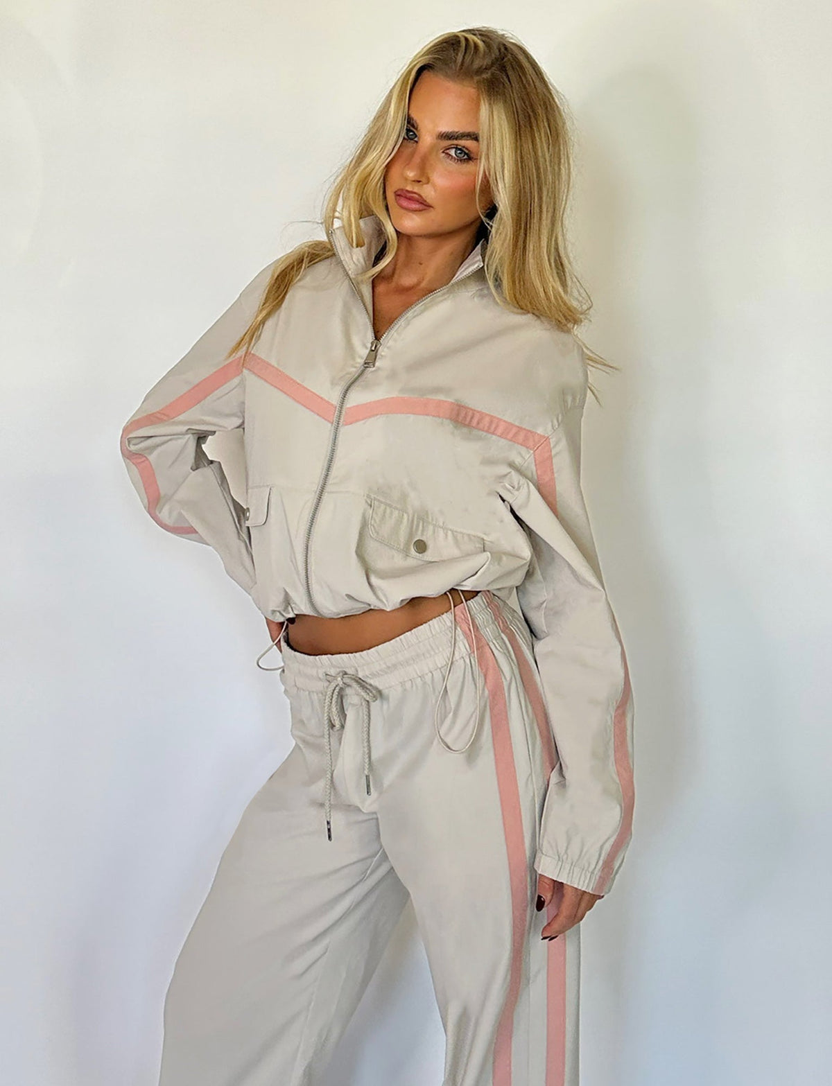 A blonde woman poses against a plain white background, showcasing the KAHANNA JACKET in Dove Grey from TIGERMIST. The jacket features a half-zip and drawstring waist, paired with pants that have an elastic waistband with a drawstring, accented by light pink stripes along the sleeves and legs.