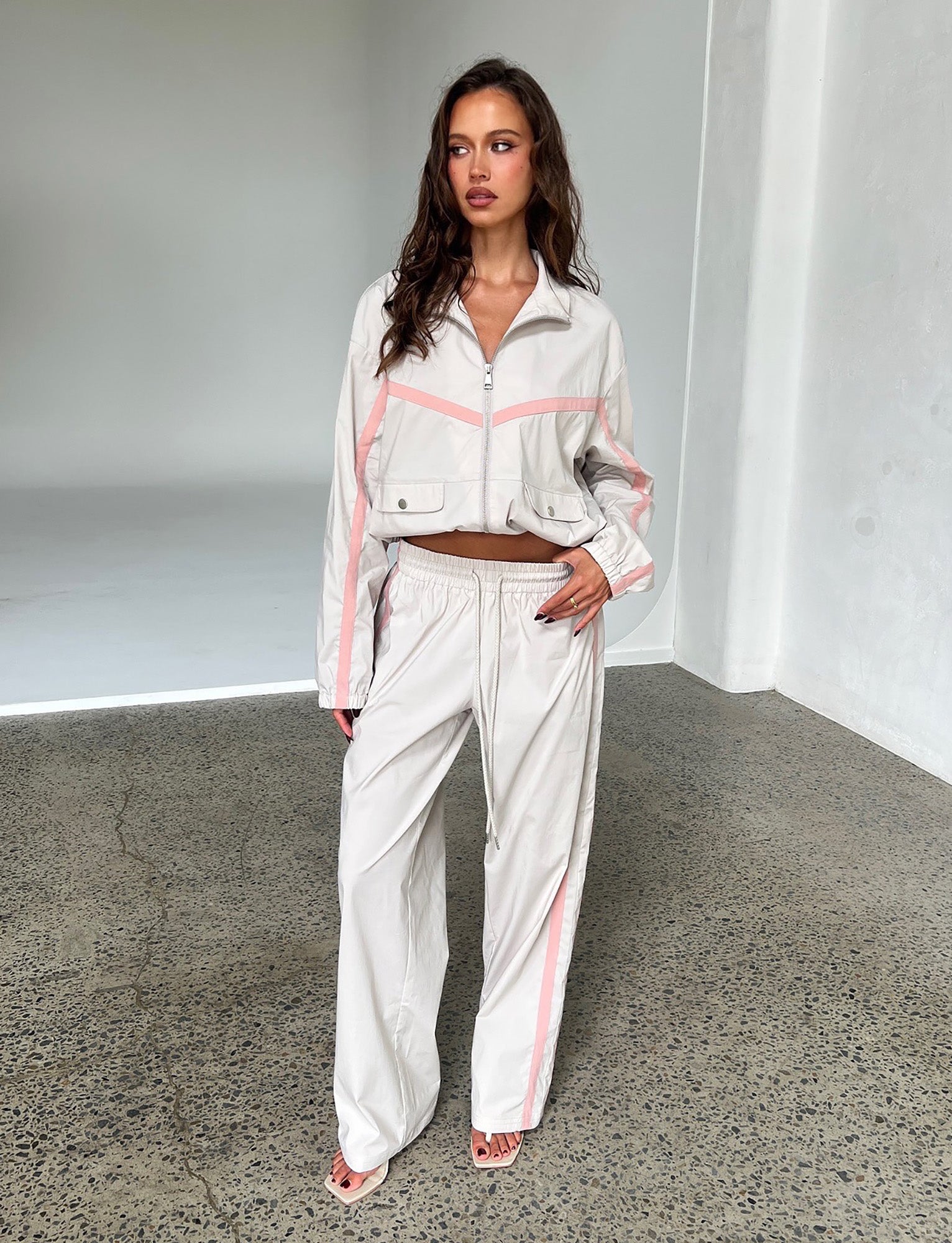 A woman stands indoors against a neutral background, dressed in a dove grey Tiger Mist tracksuit adorned with pink stripes. The outfit features a cropped jacket paired with high-waisted Kahanna pants, and she completes the look with sandals. Her long, wavy dark hair cascades down as one hand rests on her hip.