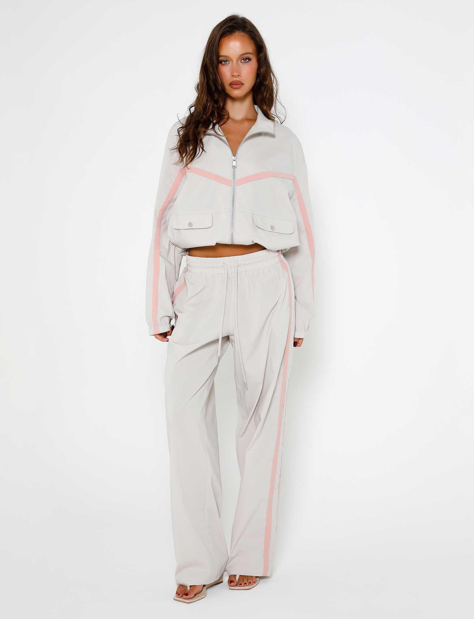 A woman stands against a plain white background, wearing the Dove Grey KAHANNA PANT from TIGERMIST, featuring pink stripes. The outfit includes a zip-up jacket and drawstring pants. She has long, wavy brown hair and is wearing heels.