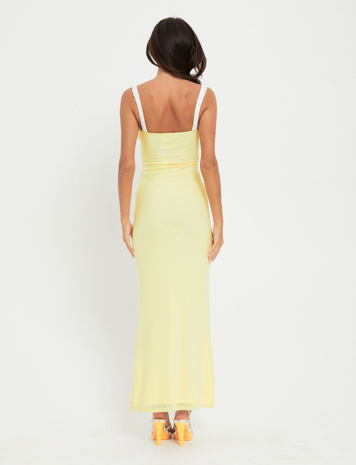 A woman with long dark hair is standing with her back to the camera, wearing an AMARAN DRESS in yellow (Lemon) from TIGERMIST. She has orange high-heeled shoes and is standing against a plain white background.