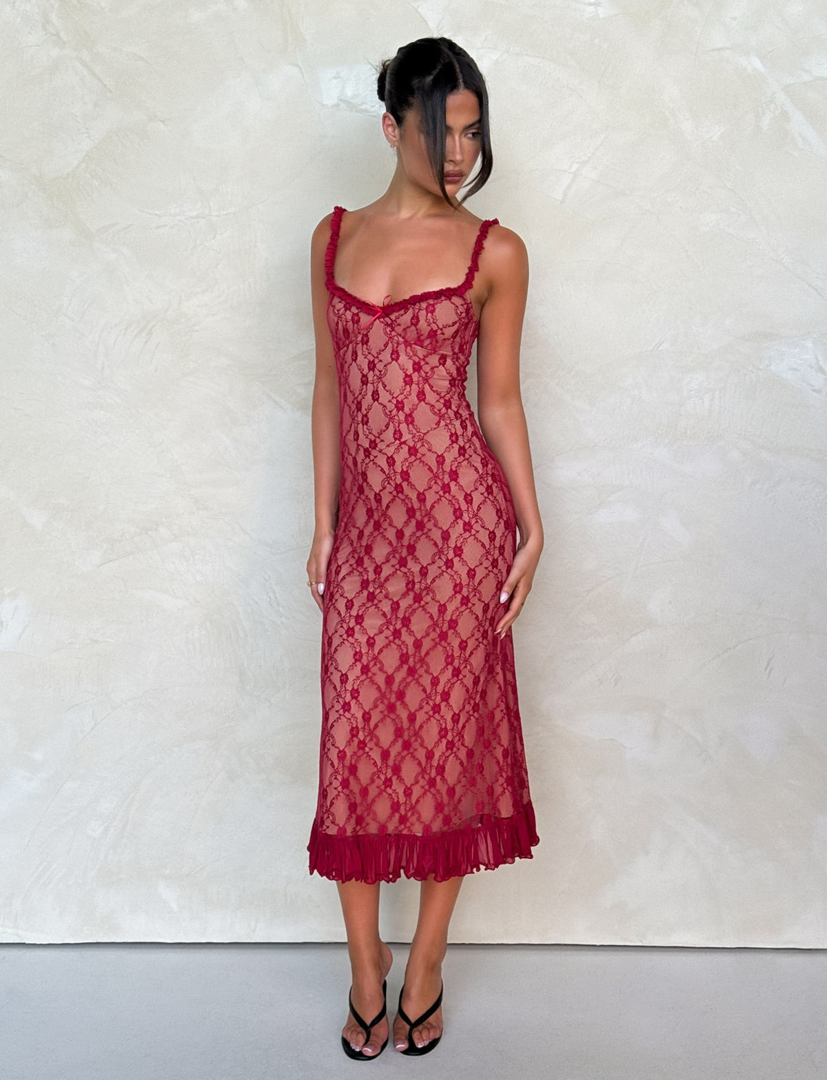 A woman in the TIGERMIST DEJA DRESS - RED, a berry red sleeveless lace midi dress with thin straps and a ruffled hem, stands by a light textured wall, looking down to her side and wearing black heeled sandals.