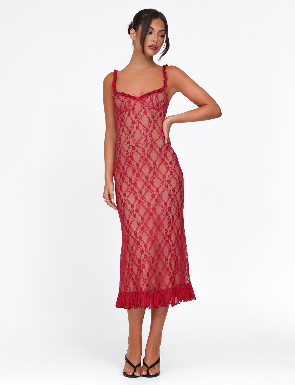 A woman poses against a white background in the TIGERMIST DEJA DRESS - RED : BERRY RED, a sheer lace dress with a fitted silhouette, ruffled hem, and thin straps, styled with black open-toe heels.