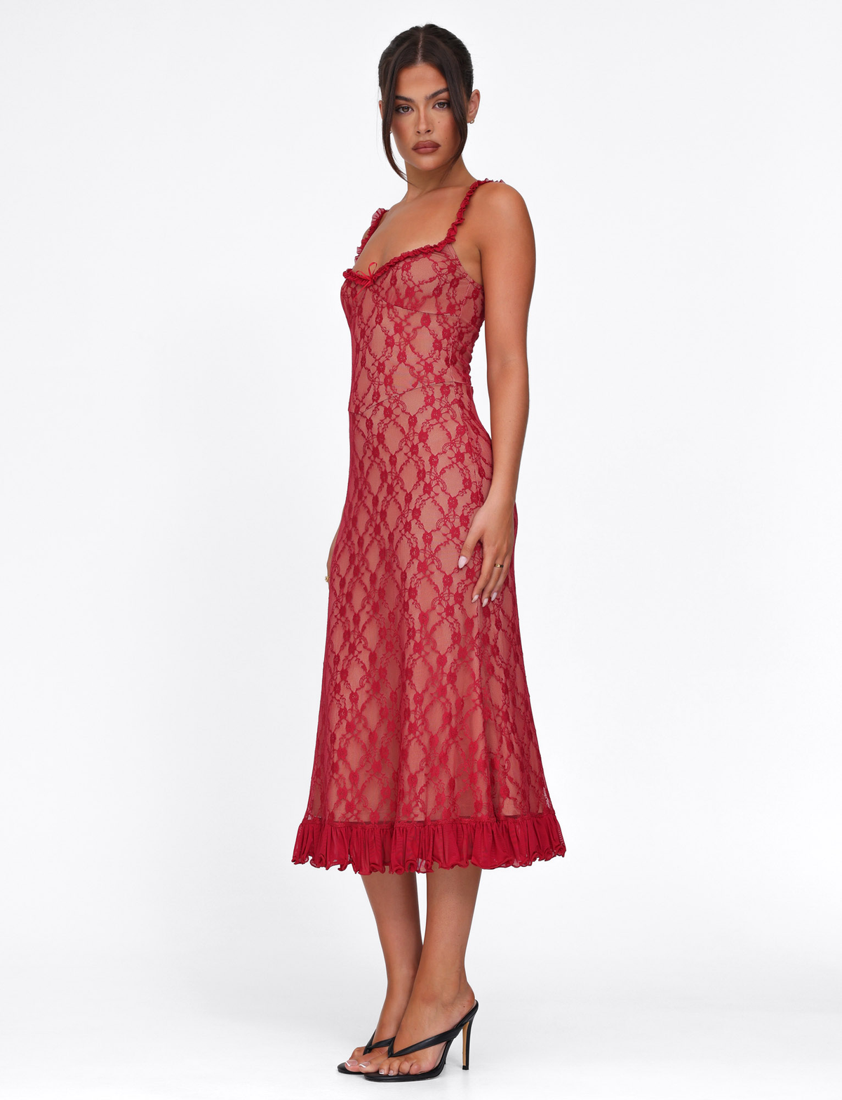 A woman stands in profile wearing the TIGERMIST DEJA DRESS - RED: BERRY RED, a fitted sheer lace dress with a ruffled hem, paired with black high-heeled sandals against a plain white background.