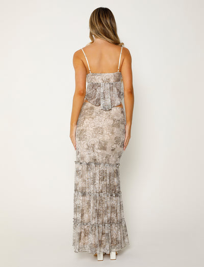 A woman with long blonde hair is seen from behind, wearing a Tigermist spaghetti strap top with a layered, ruffled back and the Alessia Skirt in Brown with a Floral Ornate pattern. The outfit features a light, neutral color palette. She is standing against a plain, light background.