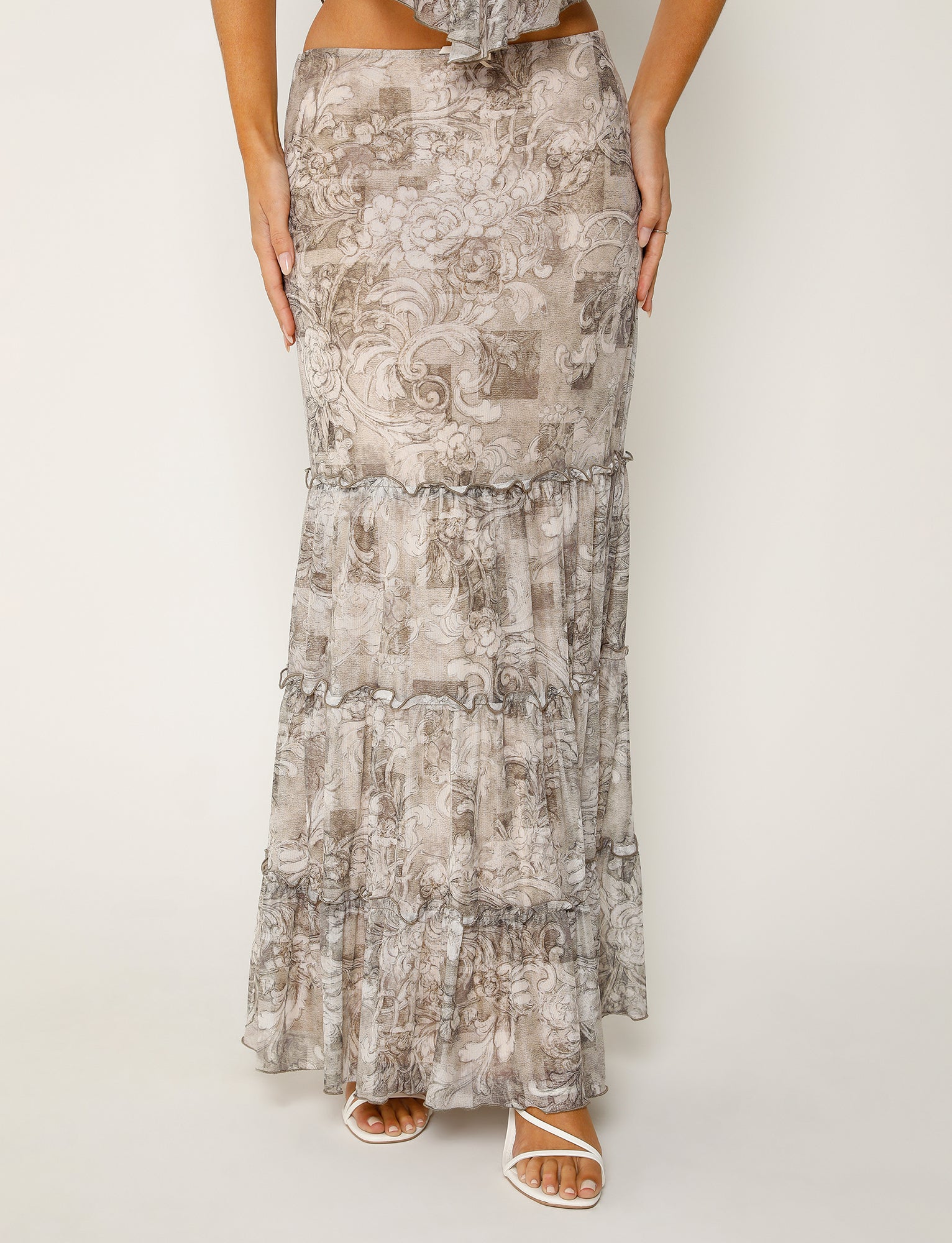 A person wearing the TIGERMIST ALESSIA SKIRT in BROWN : FLORAL ORNATE stands against a plain background. The long, flowy skirt, featuring tiered layers and adorned with an intricate gray floral pattern, reaches down to their sandal-clad feet. Their hands rest lightly on their hips.