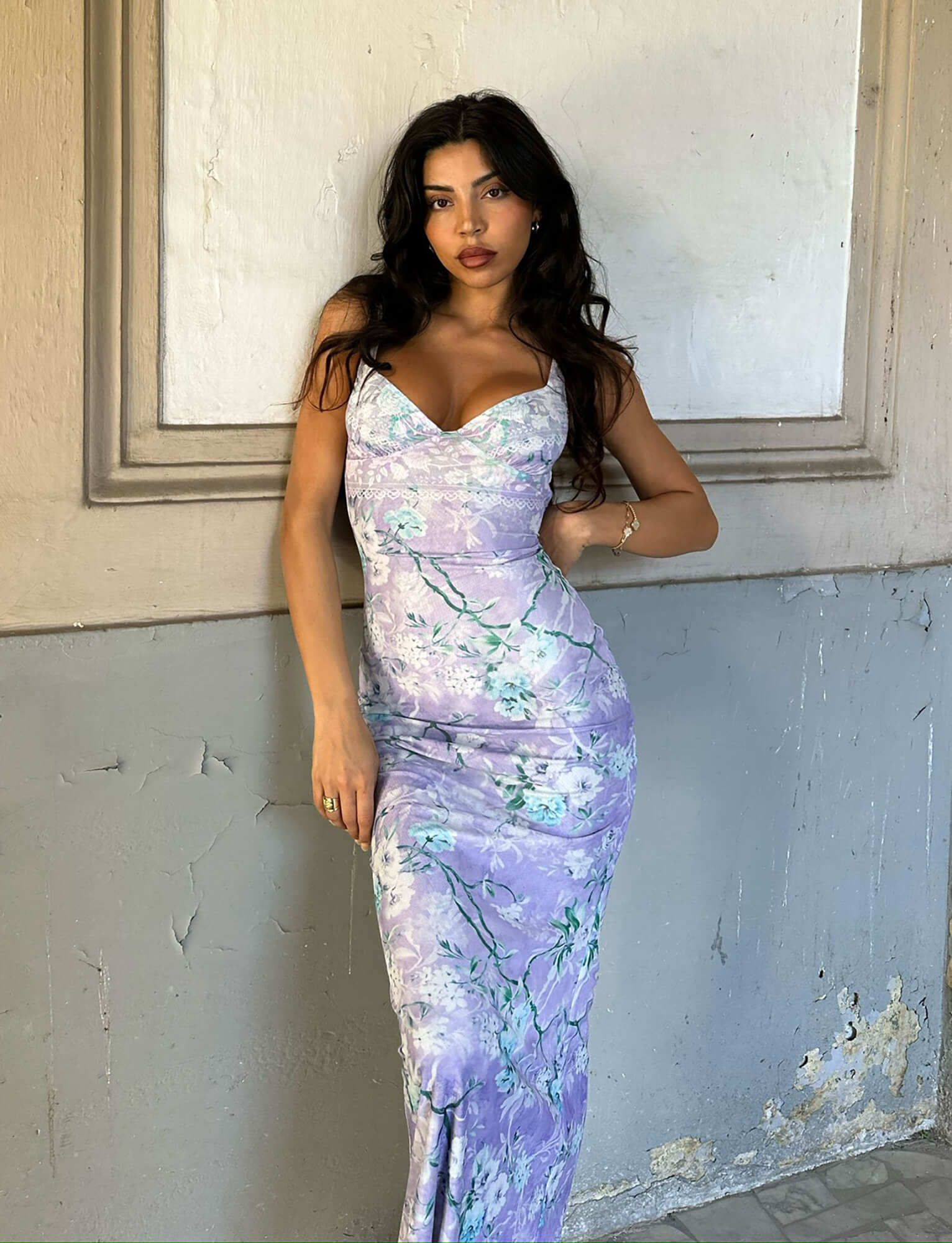 A woman with long dark hair stands against a textured wall. She is wearing the KARMEN DRESS - PURPLE : FLORAL BOARDERS from TIGERMIST, featuring a form-fitting design with a floral pattern and sweetheart neckline. Her pose is relaxed, with one hand on her hip and the other hanging by her side. The background is neutral-toned.