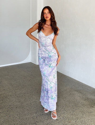 A woman poses confidently in the KARMEN DRESS - PURPLE: FLORAL BOARDERS by TIGERMIST, a fitted, sleeveless dress featuring a V-neckline and lavender floral pattern. She has long, wavy hair and stands indoors against a plain, light-colored wall. To complete her look, she wears white strappy heels from TIGERMIST.
