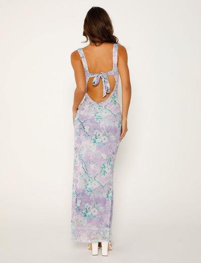 A woman with long brown hair stands facing away, showcasing the KARMEN DRESS - PURPLE : FLORAL BOARDERS by TIGERMIST, featuring an open back tied with a bow. She is wearing white platform heels and stands against a plain white background.