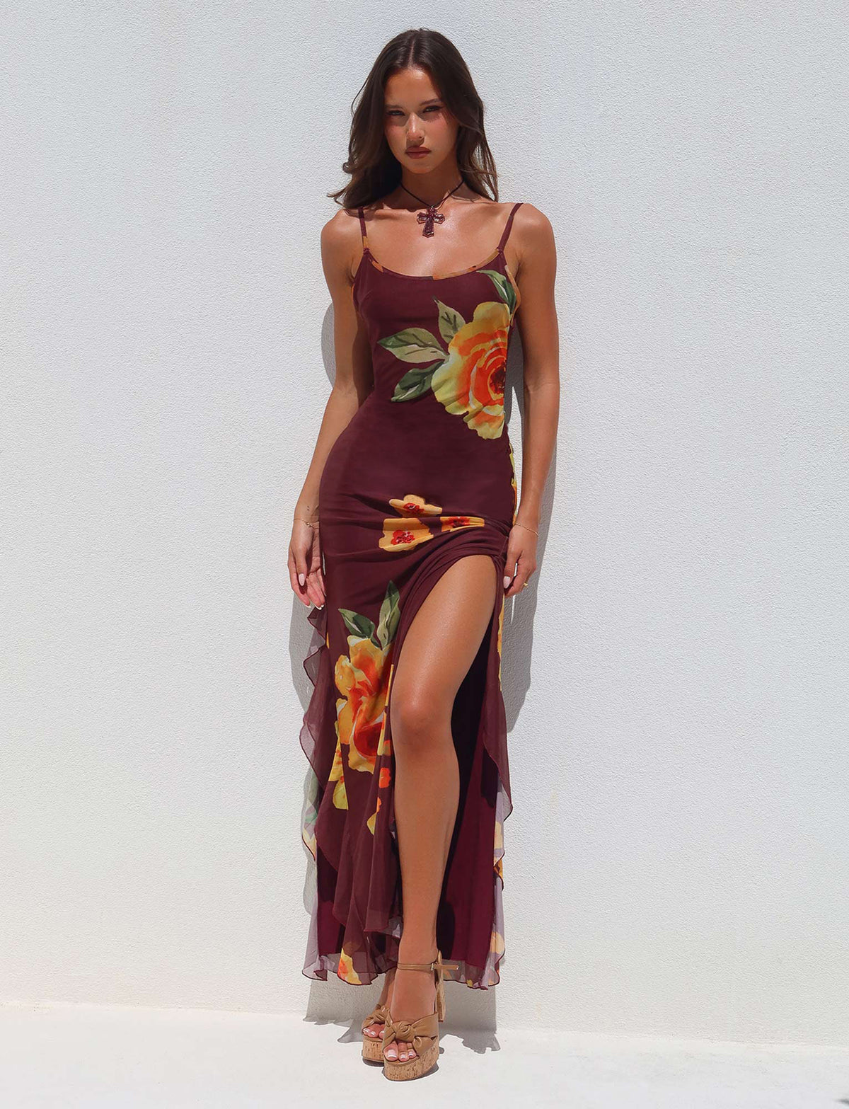 A woman stands against a white wall wearing the NEVE DRESS from TIGERMIST, a sleeveless maroon dress adorned with large yellow and orange floral prints. The dress features a thigh-high slit, and she pairs it with strappy sandals. Her long hair is styled down, and she gazes confidently at the camera.