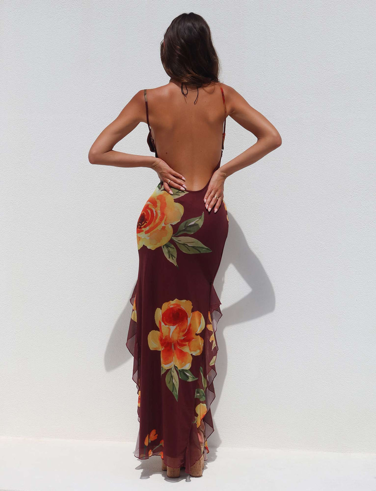 A woman with long hair stands against a plain white background, wearing a stunning Tigermist NEVE DRESS in burgundy floral. The backless dress is adorned with large, vibrant yellow and orange flowers and features delicate straps along with a flowing, layered design at the bottom. Her hands rest on her lower back.