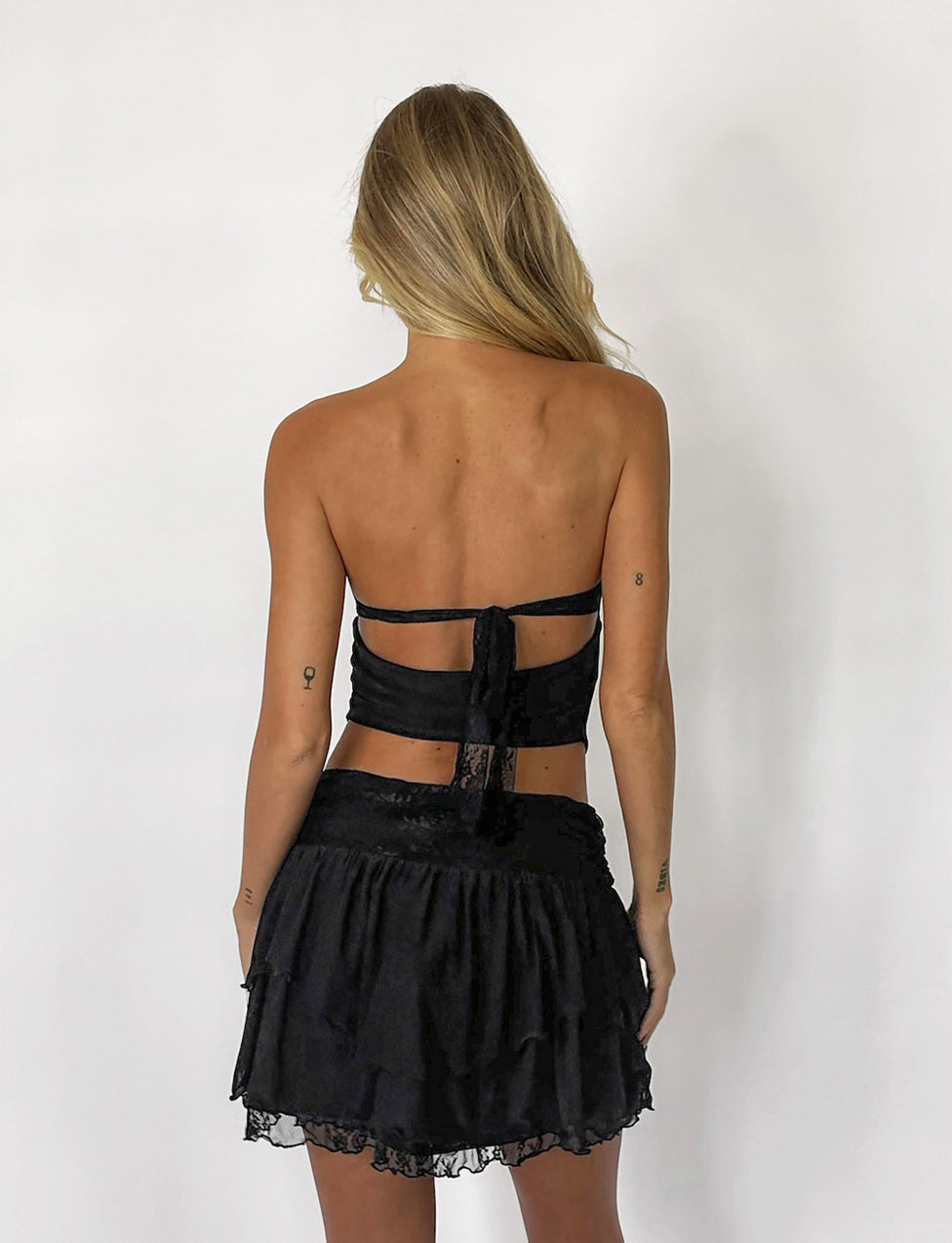 A person with long blonde hair is seen from behind, wearing the HARPER TOP - BLACK and a matching black skirt with lace trim from TIGERMIST. They have small tattoos on their arms and stand against a plain white background.