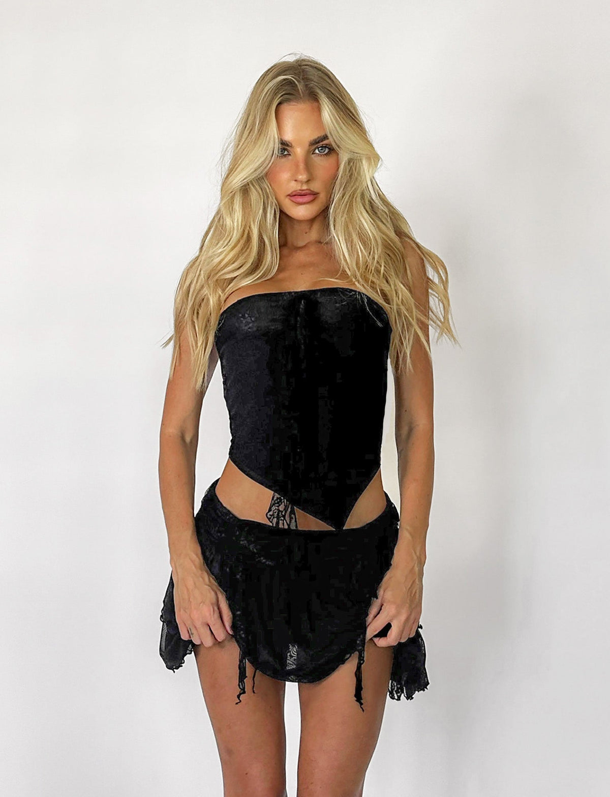A blonde woman, adorned in the elegant HARPER TOP - BLACK from TIGERMIST paired with a matching mini skirt, stands against a plain white background. Her arms rest relaxed by her sides as she gazes directly at the camera with a neutral expression.