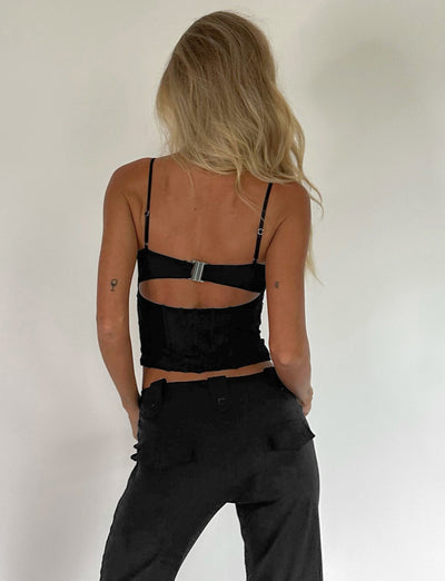 A person with long, wavy blonde hair is facing away from the camera. They're wearing a black MAIZIE TOP by TIGERMIST with a cutout at the back and black high-waisted pants. The background is plain and light-colored.