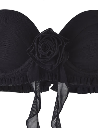 The HEAVEN TOP - BLACK from TIGERMIST is a strapless black bra adorned with a large fabric rose and flowing ribbon detail at the center. Its design showcases mesh-like fabric with ruffled trim along the bottom edge, all set against a plain white background.