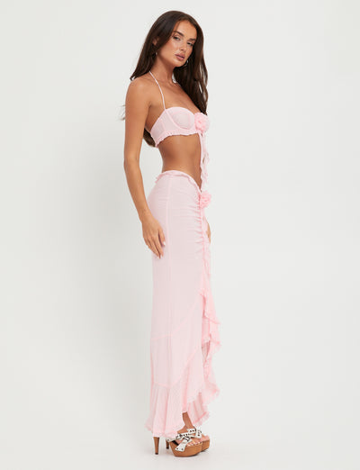 A woman with long brown hair stands sideways, looking at the camera. She is wearing the HEAVEN TOP - PINK by TIGERMIST, paired with a matching long, ruffled skirt adorned with flower-shaped decorations. She completes her look with high-heeled platform shoes. The background is plain white.