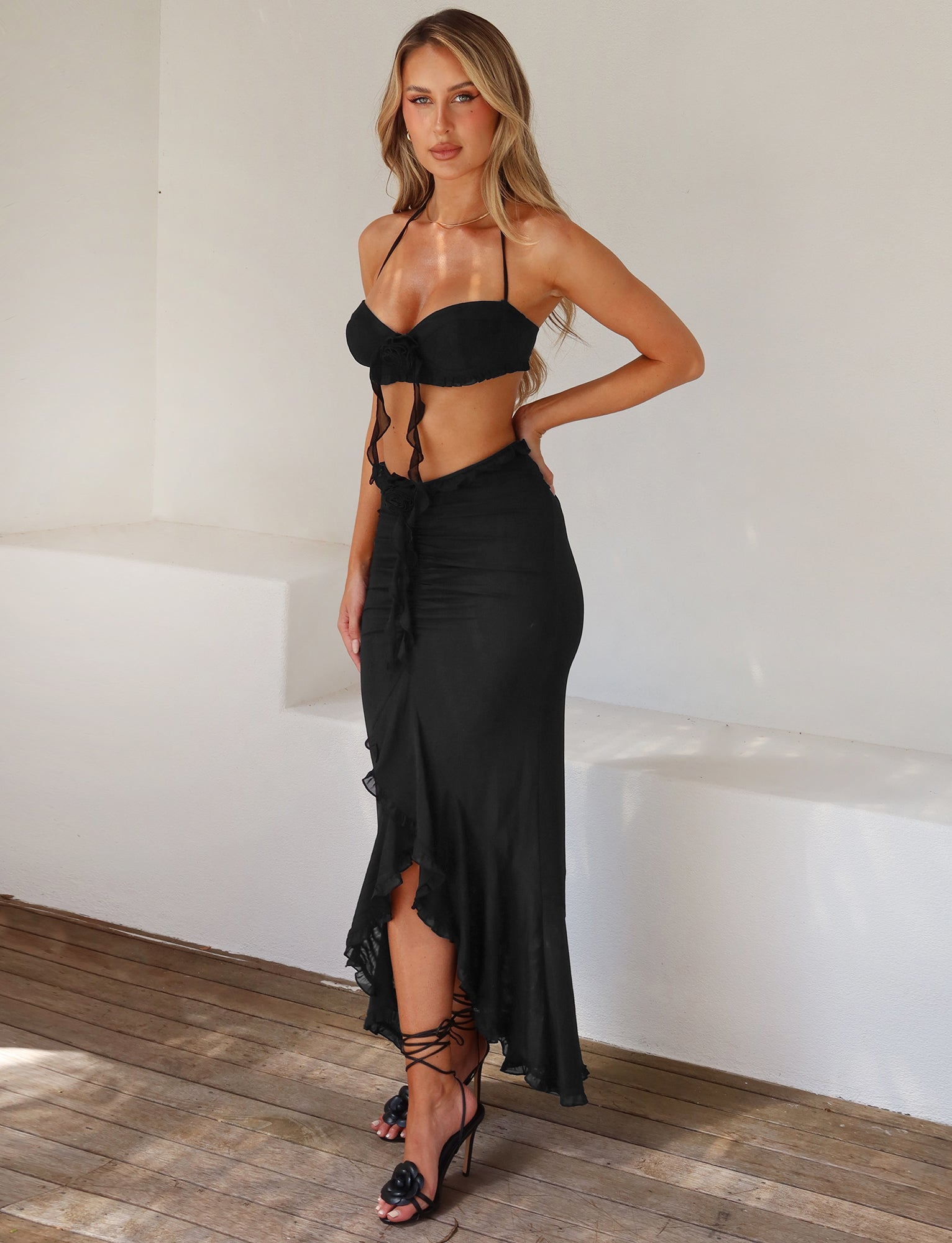 A woman with long blonde hair stands in front of a white wall, wearing a TIGERMIST black outfit featuring a bralette top with ties at the middle and the HEAVEN MAXI SKIRT - BLACK, which is high-waisted and has a ruffled hem that reaches her ankles. She completes her look with black strappy heels.