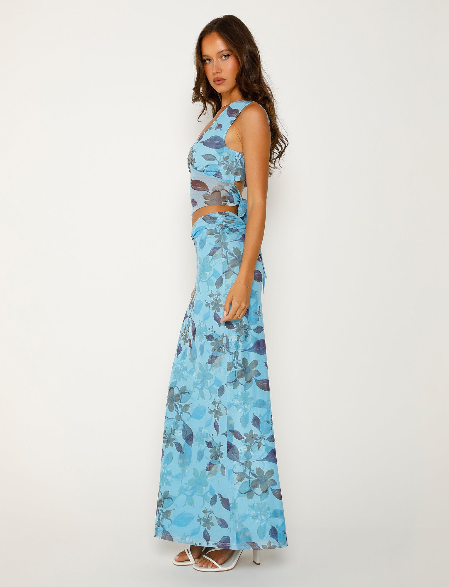 A woman with long brown hair stands sideways against a plain white background. She is wearing the MARIETTA SKIRT - BLUE : WATERCOLOUR from TIGERMIST, paired with a matching blue floral-patterned sleeveless top and white high-heeled shoes. She has a serious expression, looking directly at the camera.