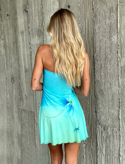 A blonde woman with long wavy hair stands with her back to the camera, showcasing a strapless, gradient mint blue Soren Dress from TIGERMIST. The background features a textured, gray wooden wall.