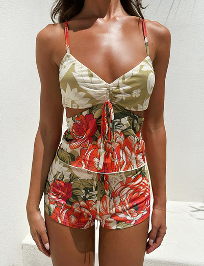 A person wearing the TIGERMIST RIOT TOP - MULTI: MACRO FLORAL: SUNSET BLOOMS two-piece outfit, which includes a sleeveless top with a drawstring and matching shorts. The fabric showcases a vibrant mix of red, orange, and white flowers with green leaves on a beige background, set against a plain white wall.