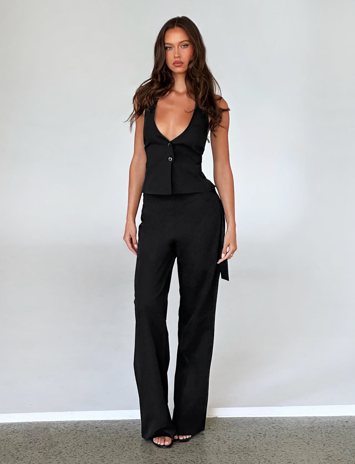 A woman with long brown hair stands against a plain white background, dressed in a chic black TIGERMIST sleeveless top with a plunging neckline and matching TRISTAN PANT - BLACK. With a neutral expression, her hands rest by her sides and her feet slightly apart on a speckled floor.