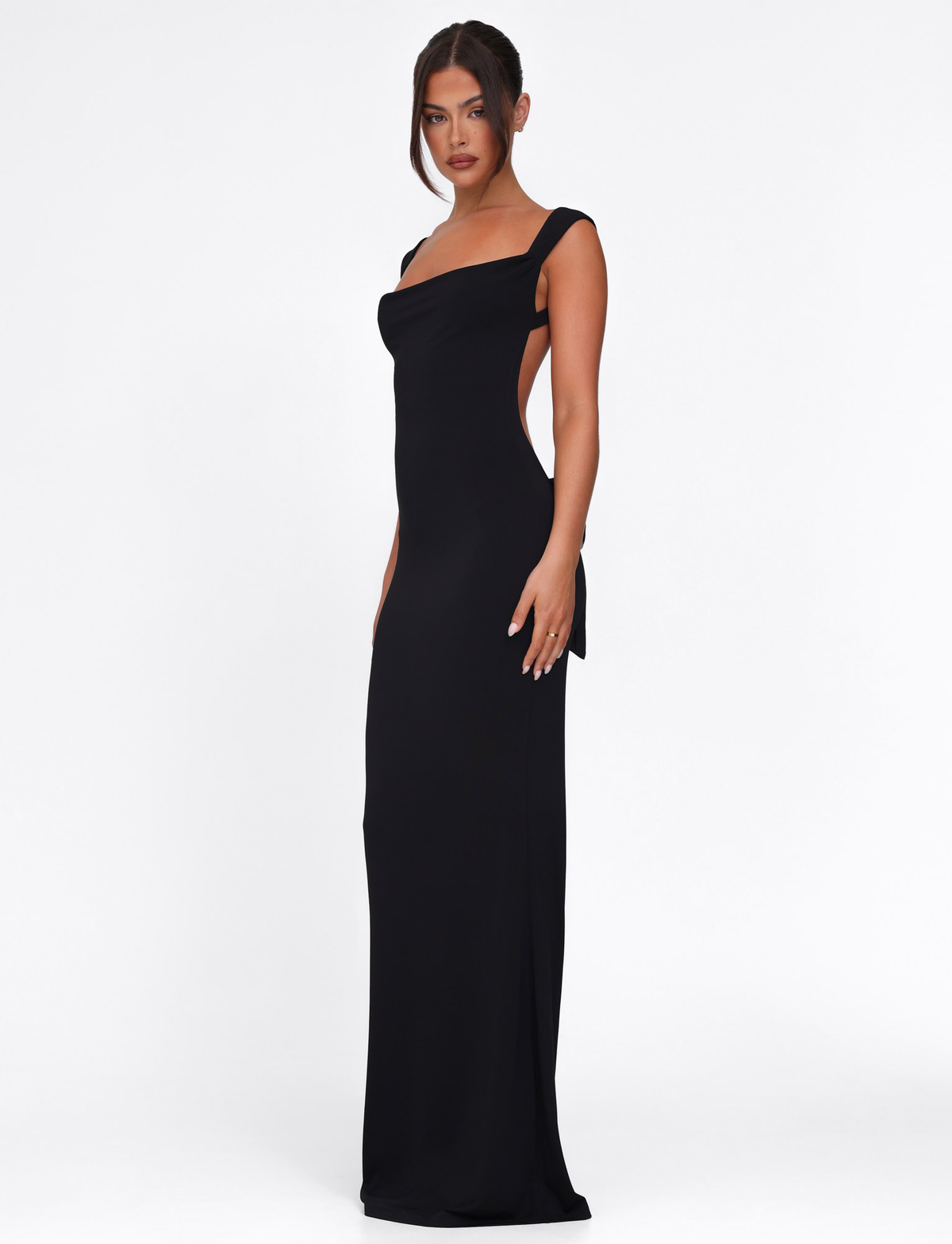 A woman models the TIGERMIST ZITA DRESS - BLACK, a black bodycon maxi dress featuring an asymmetrical neckline and open back, highlighting her silhouette against a plain white background.