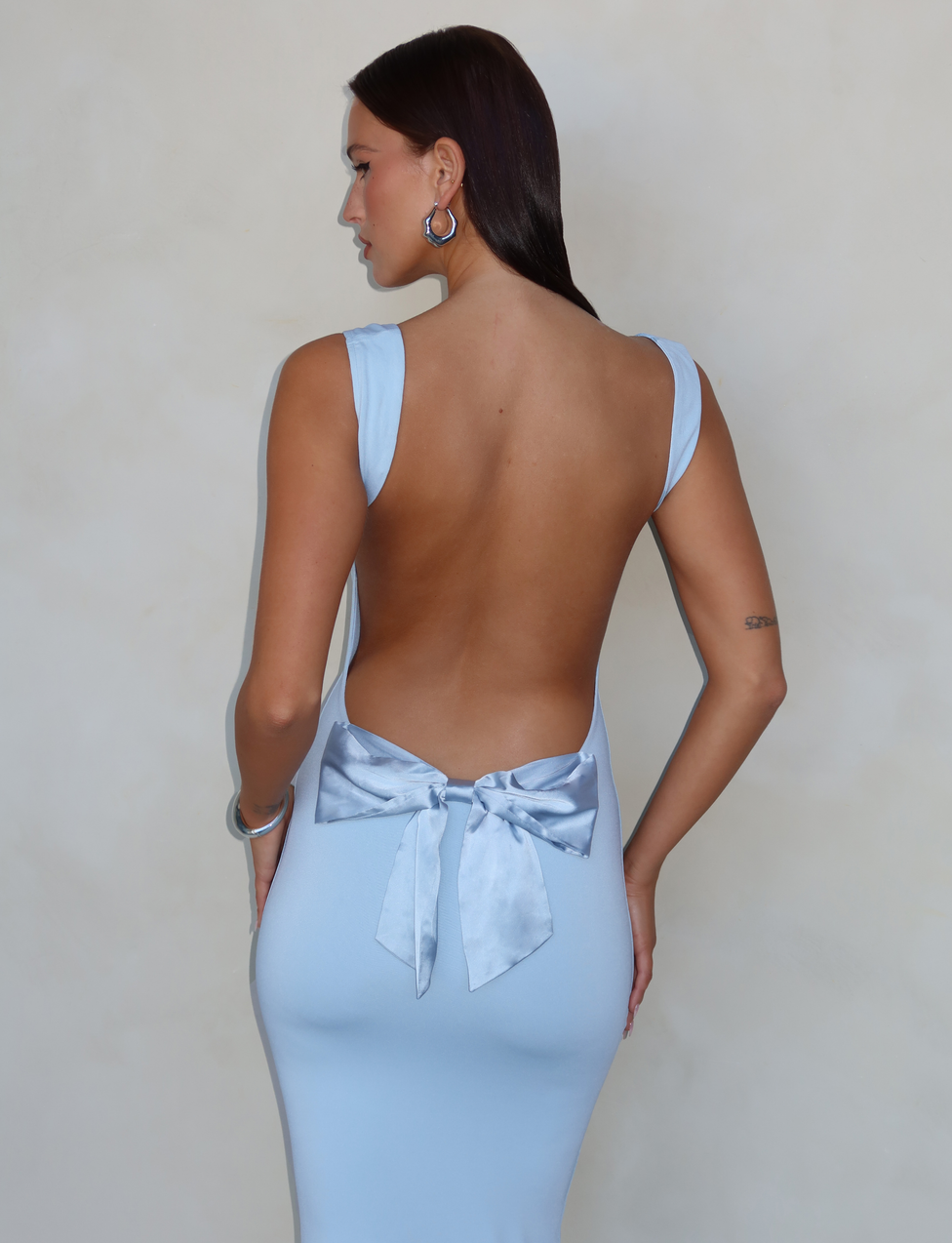 A woman faces away from the camera in the TIGERMIST ZITA DRESS - BLUE: BABY BLUE, showcasing its elegant open back with a satin bow and flattering bodycon silhouette.