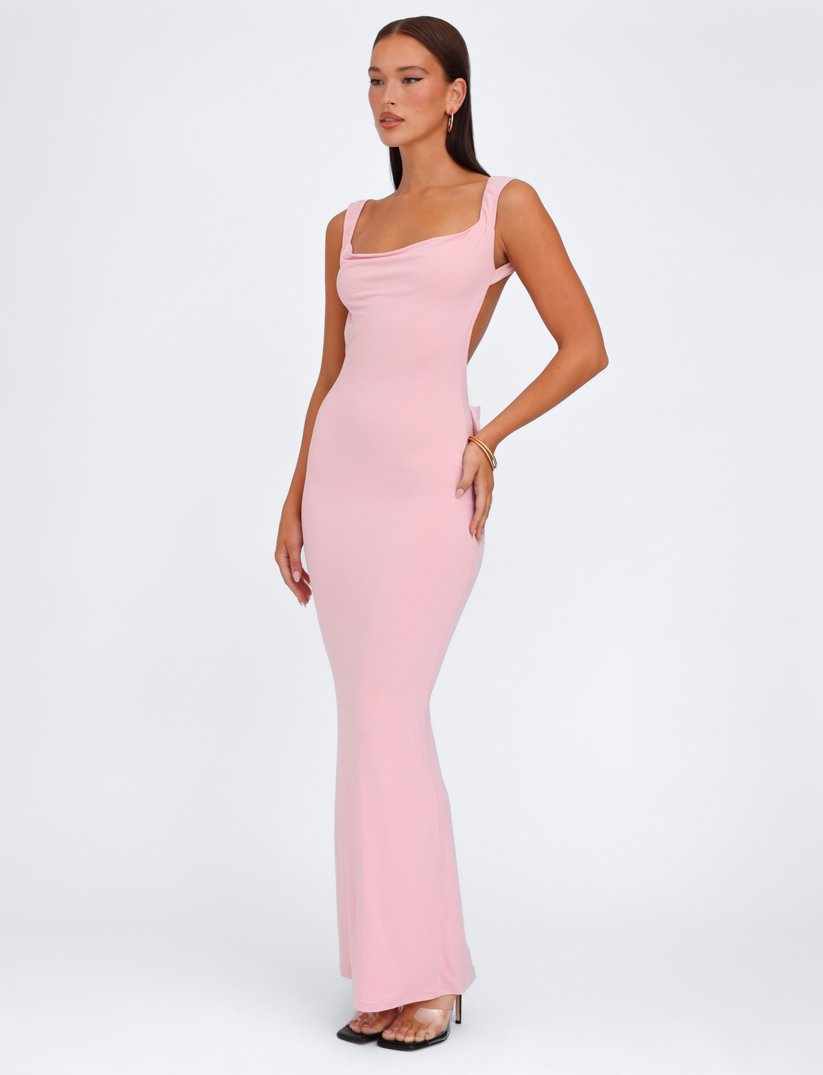 A woman poses against a white background wearing the TIGERMIST ZITA DRESS in Baby Pink—a fitted, floor-length dress with a square neckline and wide straps. She has long, straight hair, minimal jewelry, and clear heels.