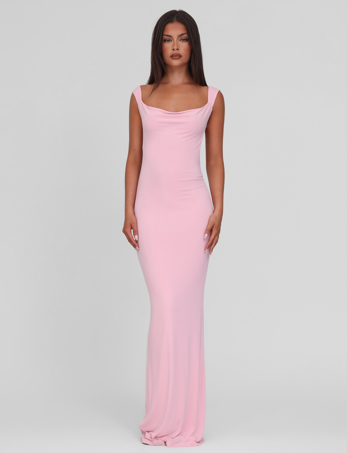 A woman with long straight brown hair stands against a light gray background, wearing the TIGERMIST ZITA DRESS in Baby Pink. This long, sleeveless bodycon dress features a draped neckline. She has a neutral expression and arms relaxed at her sides.