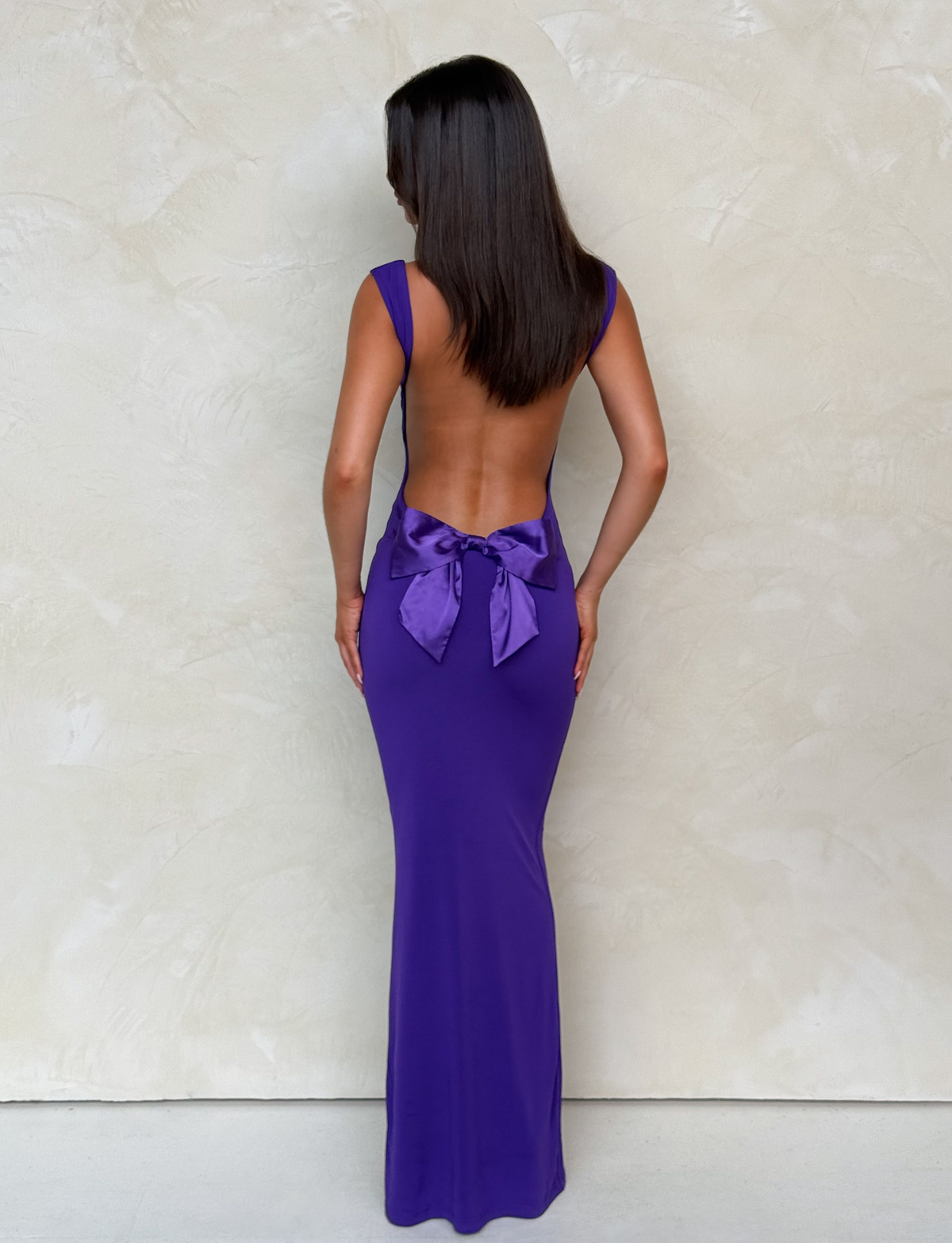 A woman stands facing a textured light beige wall, wearing the TIGERMIST ZITA MAXI DRESS in purple, featuring a large bow at the lower back. Her straight dark hair falls to her shoulders and her arms rest at her sides.