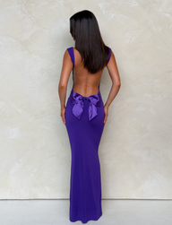 A woman stands facing a textured light beige wall, wearing the TIGERMIST ZITA MAXI DRESS in purple, featuring a large bow at the lower back. Her straight dark hair falls to her shoulders and her arms rest at her sides.