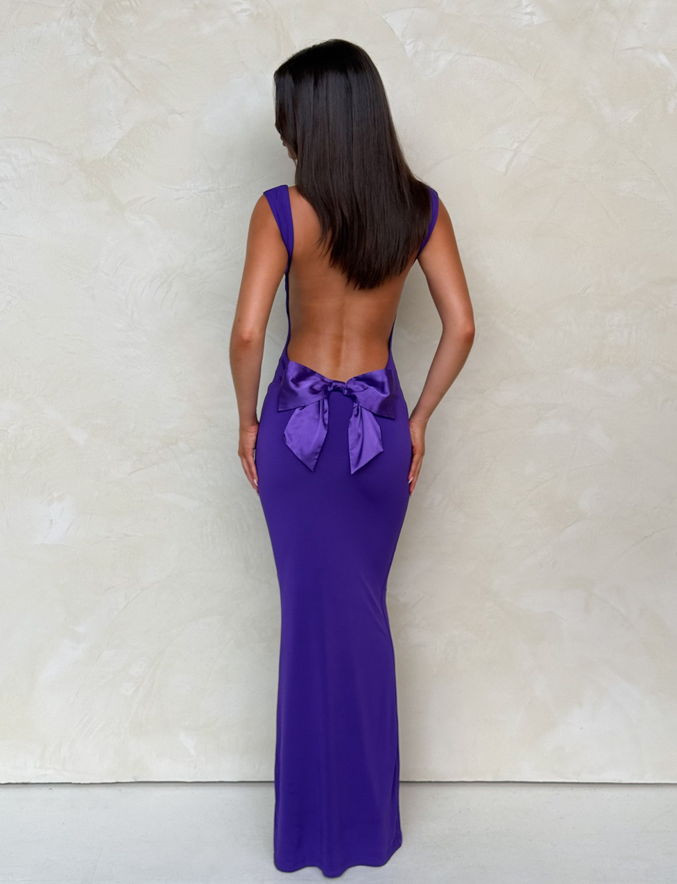 A woman stands facing a textured light beige wall, wearing the TIGERMIST ZITA MAXI DRESS in purple, featuring a large bow at the lower back. Her straight dark hair falls to her shoulders and her arms rest at her sides.